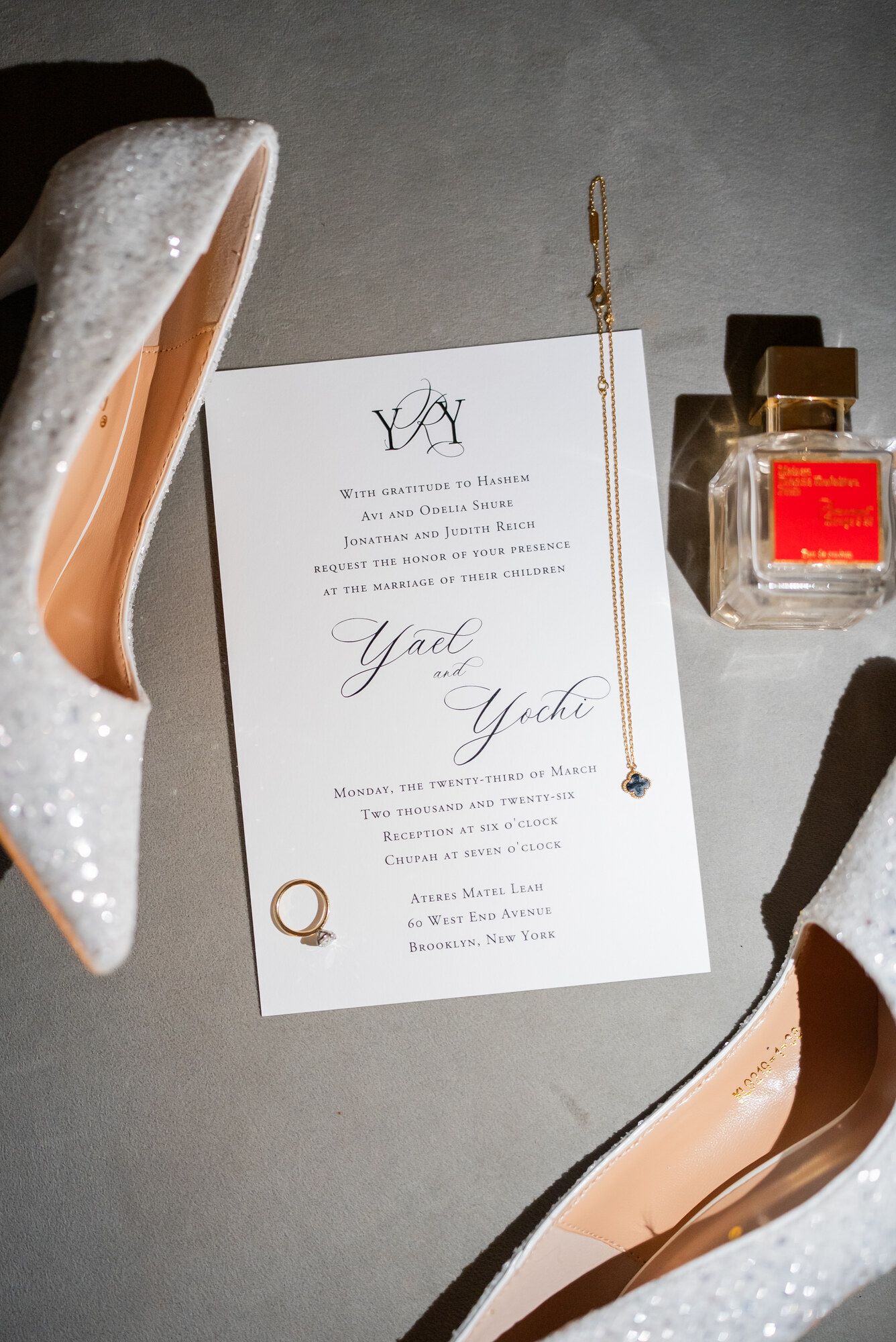 Bridal shoes, a folded wedding invitation, a gold four-leaf-clover pendant and a diamond ring arranged on grey velvet