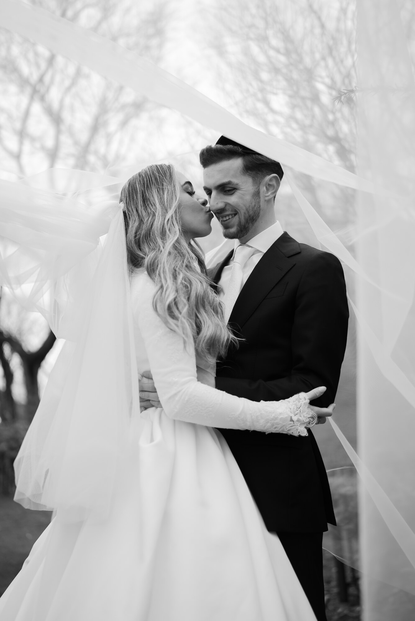 Black and white portrait of the bride kissing the groom on the nose beneath a billowing veil, bare winter trees behind them