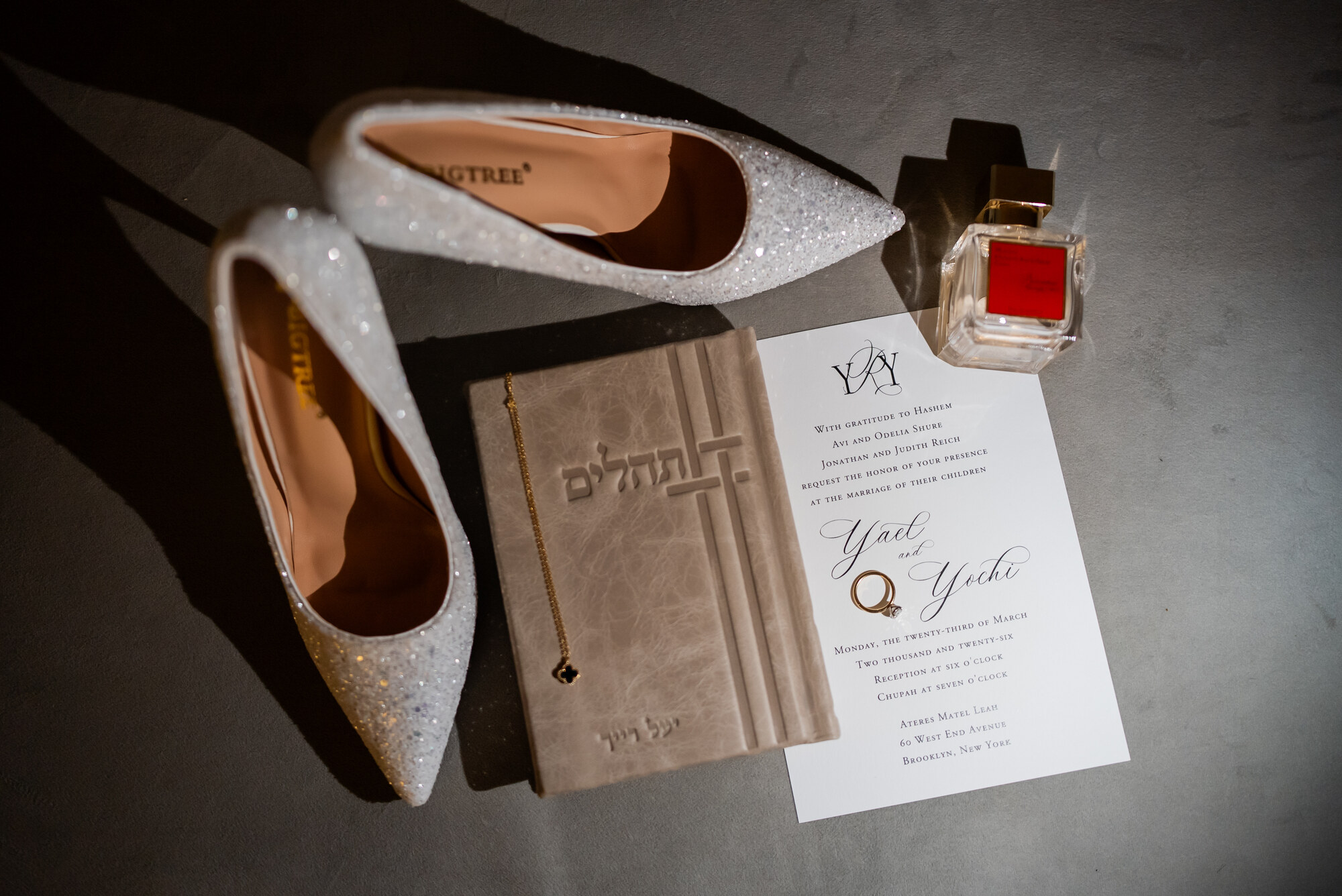 Window-lit flat lay of glittered bridal heels, an embossed leather Tehillim, a gold necklace and the wedding invitation