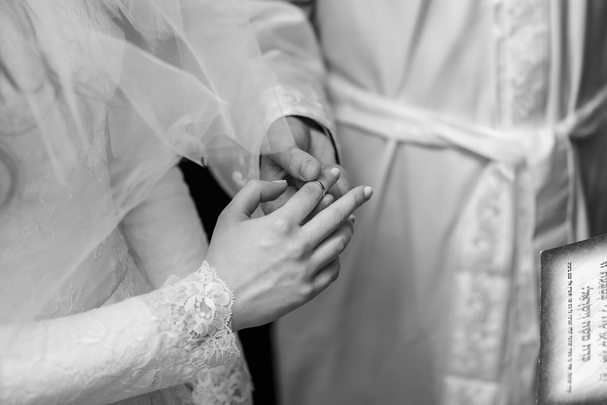 Black and white close-up of the ring being placed on the bride's finger during the ceremony, her lace sleeve and veil framing it
