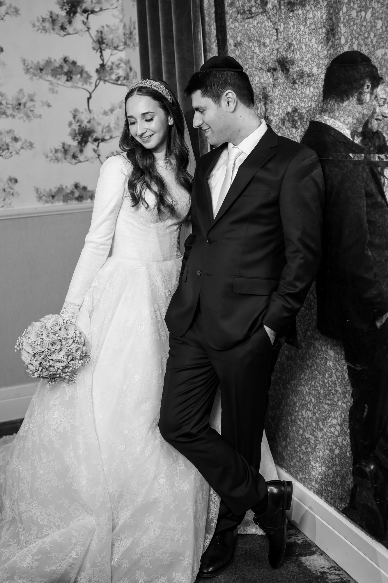 Black and white portrait of the bride and groom leaning against a patterned wall, both looking down and smiling, bride holding a white rose bouquet