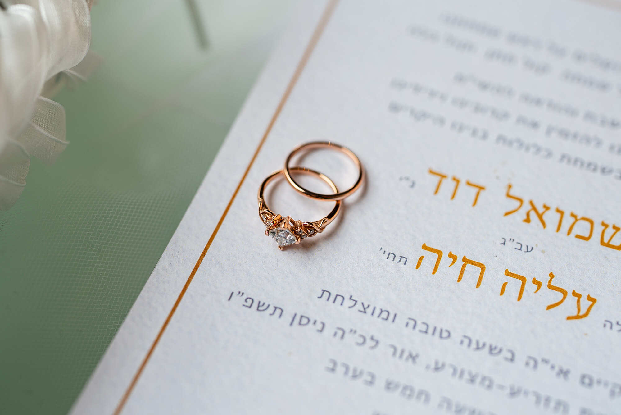 Two rose-gold wedding bands, one set with a small diamond, resting on a Hebrew wedding invitation