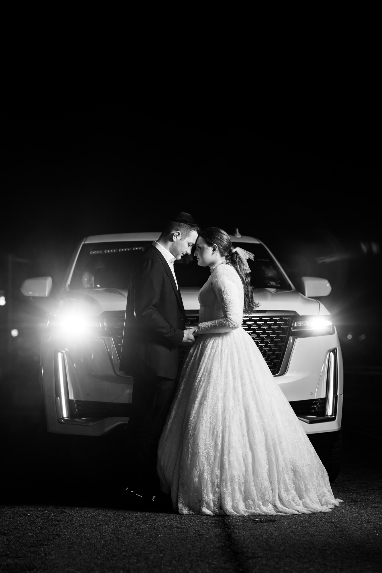 Black and white night portrait of the bride and groom forehead to forehead in front of a white SUV with its headlights on