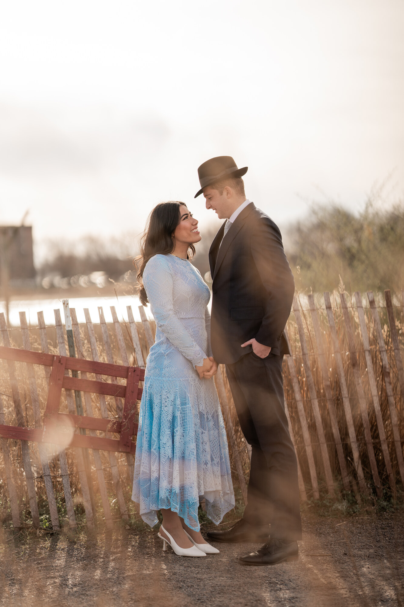 The couple facing each other beside a weathered wooden fence, she in pale blue lace and he in a dark suit and fedora, hazy golden backlight behind them