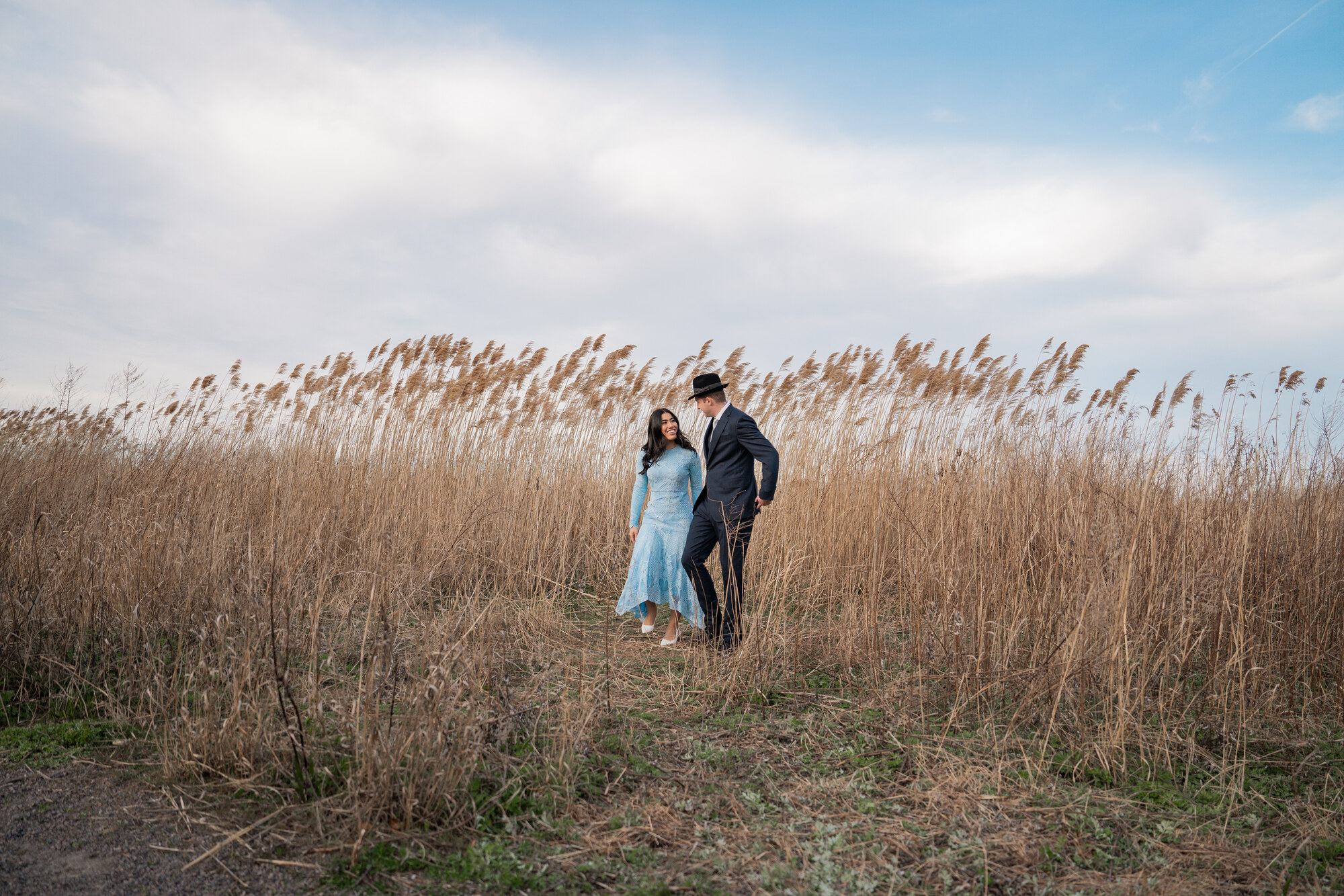The bride and groom walking through a field of tall golden reeds under a wide sky, she in pale blue lace, he in a dark suit and black fedora